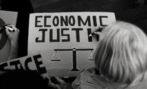 A woman writing with a black marker to create a protest sign that reads "Economic Justice"