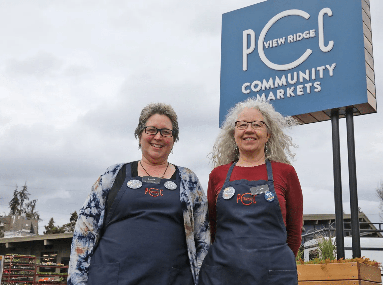 Two women wearing PCC workers aprons standing in front of a PCC grocery store sign, smiling