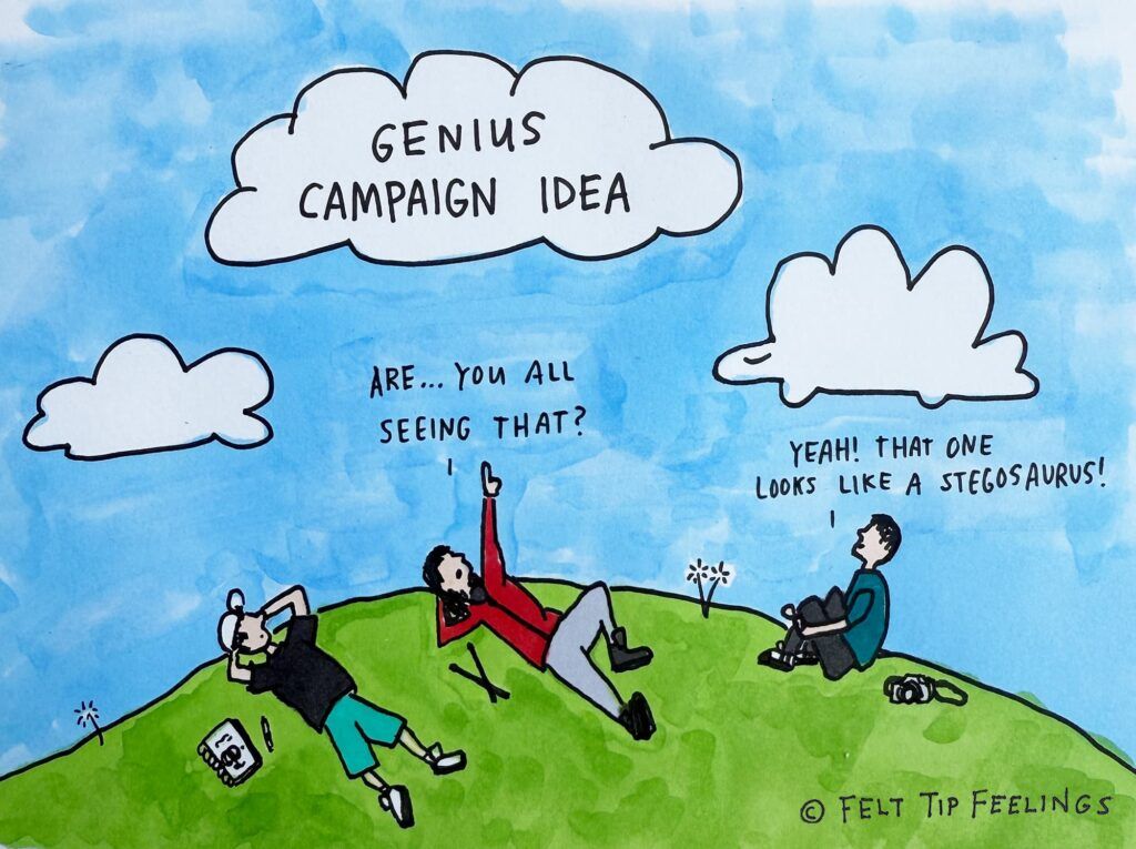 A hand-drawn cartoon of three people sitting on a grassy hill, watching clouds in a blue sky. The middle cloud is labelled "genius Campaign Idea". The person in the middle is pointing at it and saying "Are....You all seeing that?" and the person on the right is replying "Yeah! that one looks like a stegosaurus!"